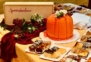 Ein Kuchenbuffet mit einer Torte in Kürbisform, vielen Kuchenstücken und einer Holzkiste mit der Aufschrift „Spendenbox“.