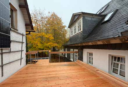 Eine neue, großzügige Holzterrasse mit Geländer an einem Haus mit Schieferdach, dahinter Bäume in Herbstfarben.