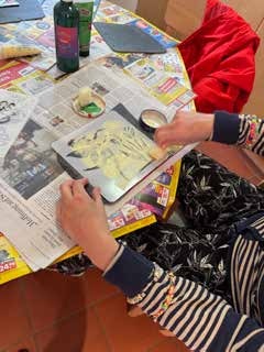 Hände malen mit gelber Farbe über eine Schablone auf einer Schiefertafel, rundherum liegen Zeitungspapier und Farbtuben.