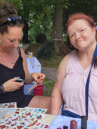 Eine Frau klebt einer lächelnden Bewohnerin mit roten Haaren ein Tattoo auf den Arm, auf dem Tisch liegen bunte Tattoo-Bögen.