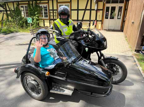 Ein Bewohner winkt aus dem Beiwagen eines schwarzen Motorrads, hinter ihm sitzt der Fahrer mit Helm und Warnweste vor einem Fachwerkhaus.